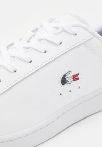 Baskets en cuir blanc avec lacets blancs et un petit logo crocodile brodé en bleu, blanc et rouge sur le côté.