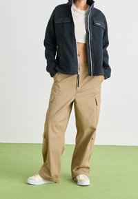 Ciemnoszara kurtka z polaru na zamek z dwiema przednimi kieszeniami, zestawiona z beżowymi spodniami typu cargo z bocznymi kieszeniami oraz kremowymi sneakersami.