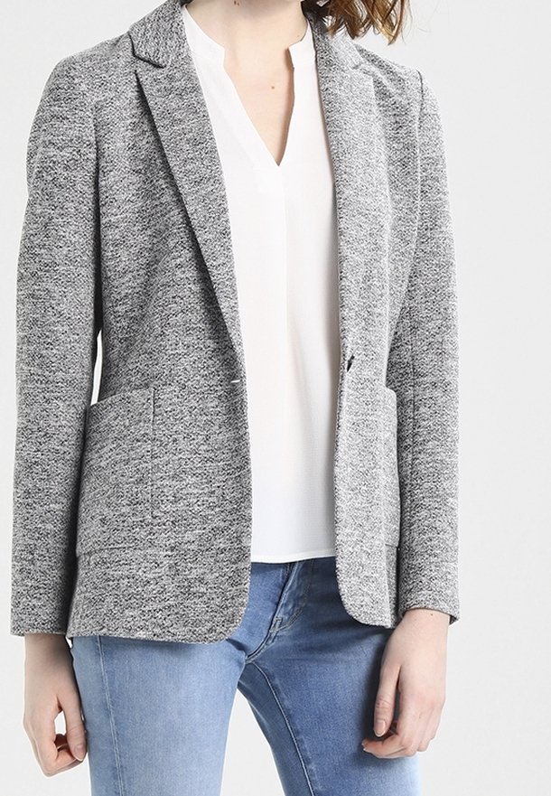 Femme portant un blazer gris texturé par-dessus un chemisier blanc à col en V et un jean bleu clair, debout devant un fond uni.