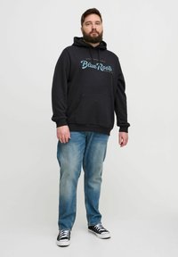Schwarzer Hoodie mit einer Fronttasche und "BlueRoots" Grafik in Türkis. Kombiniert mit hellblauen Jeans und schwarzen Sneakers mit weißen Sohlen.