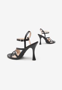 Sandalias de tacón alto negras con múltiples correas, hebilla en el tobillo, punta abierta y textura de cuero suave. Presenta un tacón afilado y una plantilla acolchada.