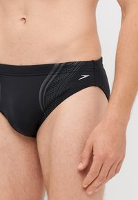 Slip da nuoto neri con un design senza cuciture, dettagli testurizzati e strisce laterali grigie con un motivo esagonale. Include un logo in argento.