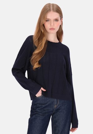 Mujer con cabello rubio largo y ondulado, vestida con un suéter de punto azul marino oscuro y jeans azules, de pie con una mano en el bolsillo sobre un fondo blanco.