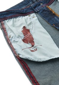 Interiorul căptușelii buzunarului din blugi albaștri cu o siluetă imprimată a unei persoane care poartă pălărie și palton deasupra textului "mcs".