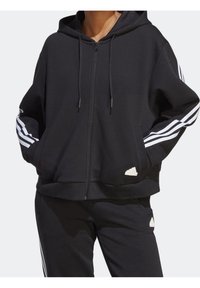 Svart zip-up hoodie i mjukt material med vita accentstripes på ärmarna, med en framficka och logodetalj vid fållen.