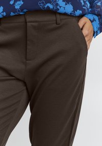 Bruna byxor med en slät yta, klassisk design, flat front och sidfickor, i kombination med en blå skjorta med blommönster.