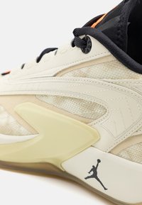 Beige sneakers med en struktureret overdel, kontrasterende sorte snørebånd og orange detaljer. Har et markant logo på hælen og en gennemsigtig sål.