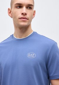 Νεαρός άνδρας με κοντά μαλλιά που φοράει μπλε μπλουζάκι EA7 Emporio Armani, κοιτάζοντας ελαφρώς προς το πλάι σε απλό φωτεινό φόντο.