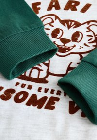 Puños acanalados verdes en una camiseta gris claro que presenta un tigre de dibujos animados marrón y texto que dice "DIVERSION CADA INCREÍBLE V." Fondo de tela texturizada.