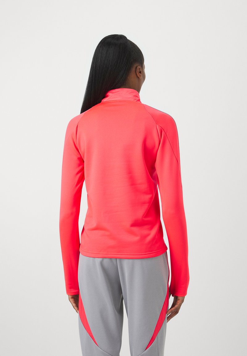 Figurbetontes, langärmliges Sportoberteil in leuchtendem Pink, mit hohem Kragen und glatter Textur, kombiniert mit grauen Hosen mit roten Akzenten.