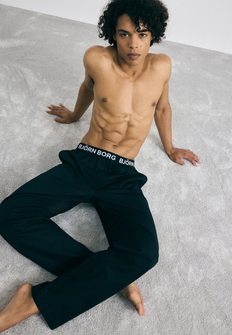 Fekete pizsamanadrág fehér "BJÖRN BORG" derékpánttal, könnyű anyagból, laza szabással, mezítláb egy texturált szürke felületen.