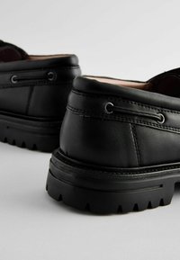Zapatos de cuero negro con superficie texturizada, costuras decorativas y ojales circulares. Suela de goma gruesa con un patrón de relieve robusto.