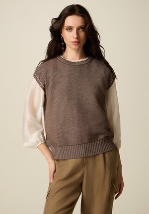 Mujer con cabello largo y oscuro, vistiendo un suéter marrón de punto sin mangas sobre una blusa blanca de mangas abullonadas y pantalones cargo color beige, de pie frente a un fondo liso.