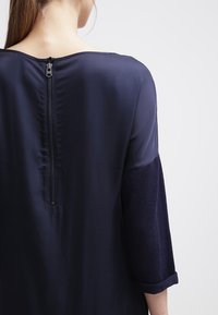 Abito blu navy con maniche a ¾, texture in satin e chiusura con zip visibile sul retro. Presenta un pannello di tessuto a contrasto sulle maniche.