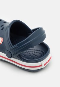 Sabots Crocs bleu marine avec sangle de talon réglable, bande de semelle blanche et rouge, semelle intérieure texturée et trous de ventilation.