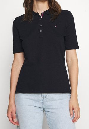 Frau trägt ein schwarzes Poloshirt mit kurzen Ärmeln und Knöpfen und hellblaue Jeans, steht vor einem schlichten weißen Hintergrund.