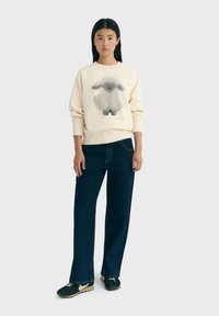 Giovane donna in piedi, che indossa un pullover crema con un'illustrazione di pecora nera, jeans blu scuro e sneakers nere su uno sfondo neutro.