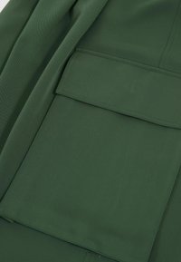 Grön tyg med en slät textur som har en stor, kvadratisk ficka. Materialet verkar hållbart med ett subtilt fiskbensmönster.