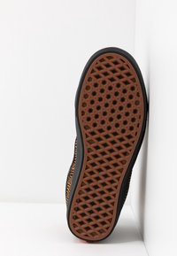 Semelle de chaussure avec empreinte en caoutchouc marron présentant des motifs hexagonaux et en forme de losange, des bords noirs et une partie supérieure noire et marron tachetée.