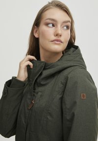 Dark green hooded jacket featuring a textured fabric, elastic cuffs, and a brown leather accent on the front for closure.