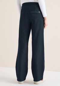 Femme portant un pantalon large bleu marine foncé avec poches arrière, associé à des baskets blanches et un haut à manches longues blanc, posant devant un fond beige.