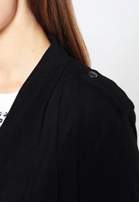 Trench coat in suede nero con colletto a revers, dettagli con bottoni sulla spalla e texture liscia. Vista ravvicinata che mostra i dettagli del tessuto.