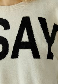 Cream sweater featuring large, textured black letters spelling "SAY" across the front. Ribbed neckline adds detail.