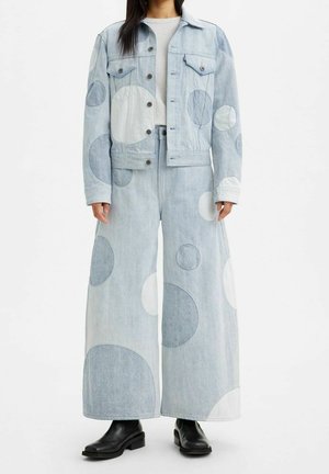 La personne porte une veste en jean bleu clair et un jean à jambes larges avec de grandes patches circulaires de différentes nuances, associée à des bottines noires.
