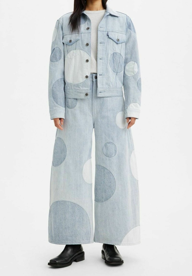 La personne porte une veste en jean bleu clair et un jean à jambes larges avec de grandes patches circulaires de différentes nuances, associée à des bottines noires.
