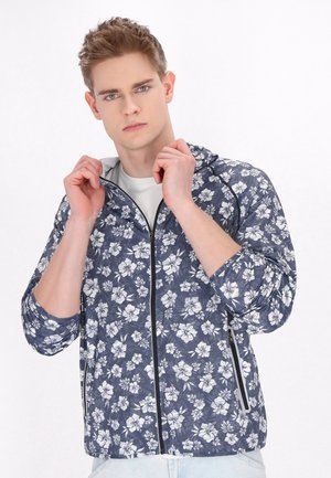 Blomster-zip-jakke i mørkeblå med hvidt blomster mønster. Har hætte, lange ærmer og sidelommer med lynlåse. Glat tekstur.