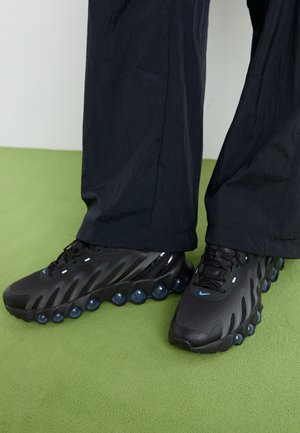 Pies con zapatos deportivos negros con suelas acolchadas de aire azul y pantalones negros, sobre una alfombra verde.