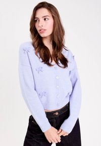 Ung kvinde med langt brunt hår, iført en lys lilla knap cardigan med små sløjfedetaljer og sorte højtaljede jeans.