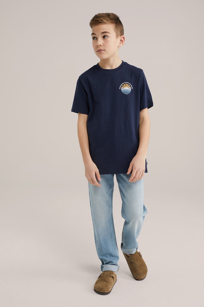 Granatowa bawełniana koszulka z okrągłym dekoltem i krótkimi rękawami, z wielokolorowym wzorem słońca i fal na piersi. Jasne dżinsy i brązowe drewniaki.