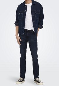 Mörk denimjacka med två bröstfickor, bärs över en vit t-shirt. Mörkblå slim-fit jeans och svarta låga sneakers kompletterar outfiten.