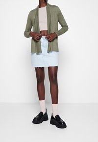 Vihreä oliivinvärinen neuletakki pitkillä hihoilla, kerroksittain vaaleanbeigea crop-topin päällä. Vaaleansiniset farkkuhameet, joissa on reviirihelma, yhdistetty mustiin korokepolkimiin.