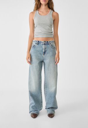 Kobieta w jasnoszarej bezrękawnikowej skórzanej bluzce, szerokich spodniach dżinsowych w wyblakłym niebieskim kolorze oraz brązowych szpiczastych butach, stojąca na jednolitym tle.