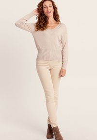 Beige gestrickter Pullover mit V-Ausschnitt und geripptem Saum, kombiniert mit hellbeigen Slim-Fit-Hosen und braunen Ankle Boots, die ein strukturiertes Design präsentieren.