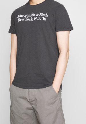 Man wearing a black Abercrombie & Fitch t-shirt with white text and gray pants, hands in pockets against a plain background.
