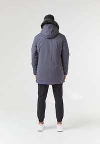 Šedá parka s kapucí podšitou kožešinou, s rovný střih. Spárována s černými joggingovými kalhotami a bílými teniskami. Jednoduché pozadí.