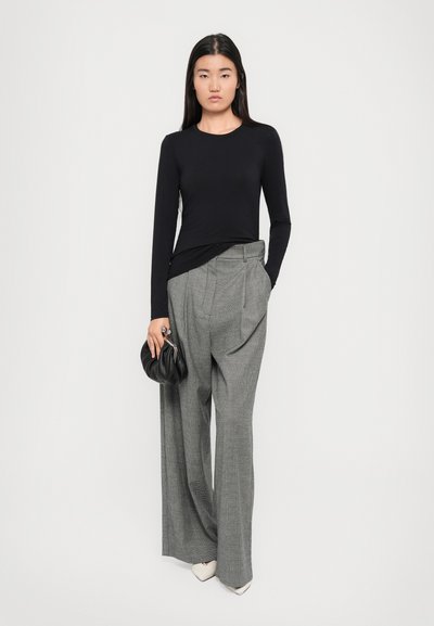 Un haut noir à manches longues associé à un pantalon large gris à carreaux taille haute. Les accessoires incluent une pochette noire et des chaussures blanches à bouts pointus.