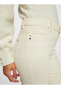 Vaqueros de talle alto en color beige, confeccionados en tela suave, con cinco bolsillos, detalles en metal plateado y costuras sutiles en los laterales.