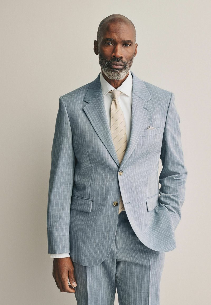 Costume bleu clair à rayures avec une coupe ajustée, veste à un bouton, revers cranté, et cravate beige, avec un pochette.