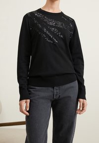 Schwarzer Pullover mit langen Ärmeln und einem diagonalen Paillettenmuster. Hergestellt aus weichem Stoff, mit einem gerippten Ausschnitt und Bund. Kombiniert mit dunkler Jeanshose.