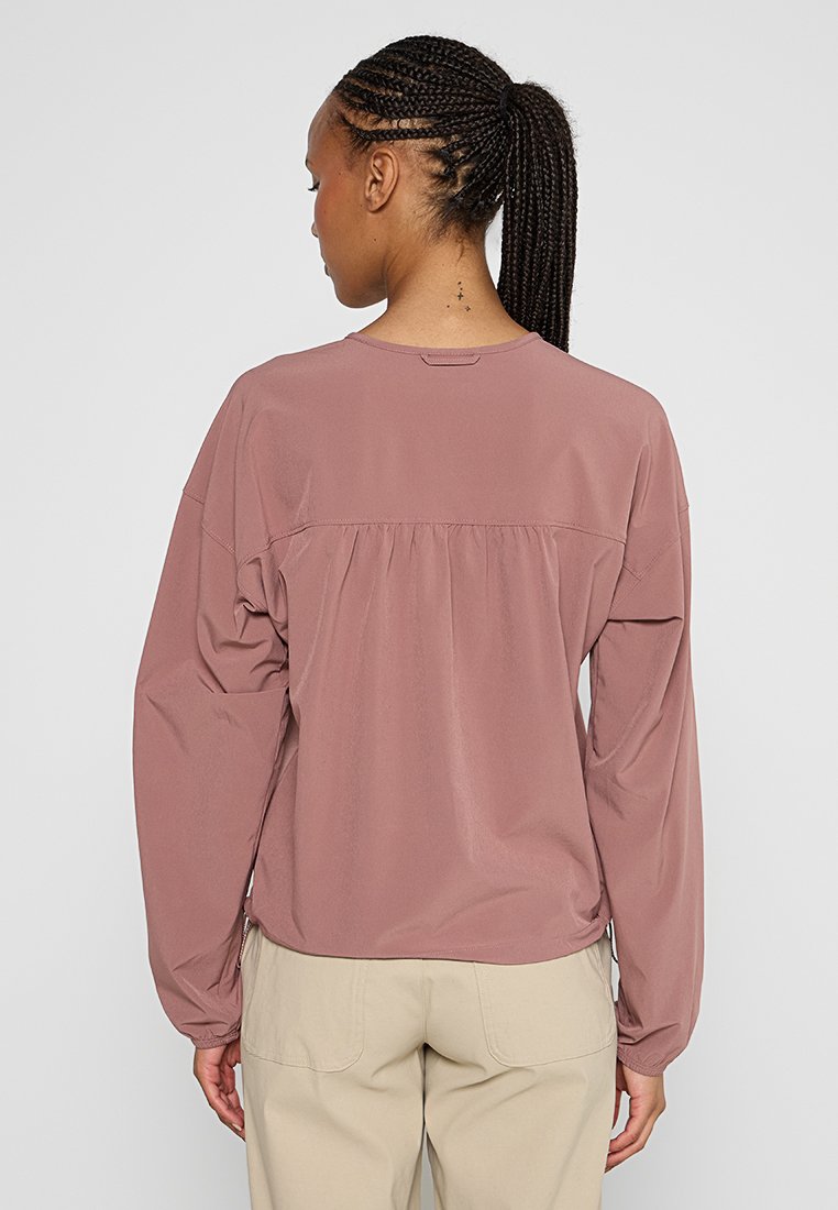 Koszula z długim rękawem w kolorze mauve, wykonana z gładkiego materiału. Ma luźny krój z marszczoną karczkiem z tyłu i zaokrąglonym dołem, zestawiona z jasnymi spodniami.