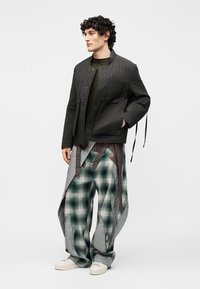 Νέος άνδρας φοράει μαύρο μπουφάν με κορδόνια, φαρδιά παντελόνια σε πράσινο και γκρι καρό με λεπτομέρειες δεσίματος, και λευκά αθλητικά παπούτσια, στέκεται αριστερά.