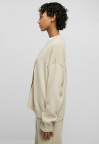 Beige oversized cardigan with a V-neck and dropped shoulders. Textured knit material, ribbed cuffs, and a relaxed fit.