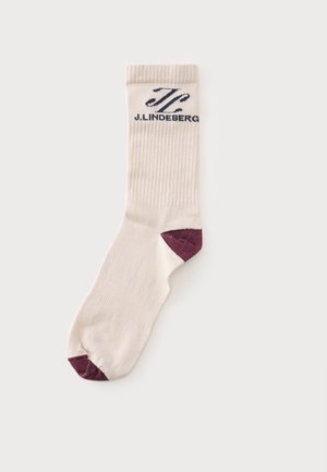 Chaussette beige mi-haute avec partie supérieure côtelée, accents violet foncé sur la pointe et le talon, et logo J. Lindeberg noir près du bord.