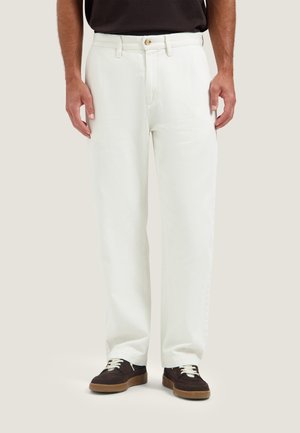 Uomo che indossa pantaloni bianchi a gamba dritta, camicia nera e sneakers nere con lacci bianchi, in piedi davanti a uno sfondo semplice.