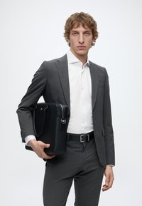 Jeune homme en costume gris et chemise blanche tenant une mallette en cuir noir sur un fond clair uni.