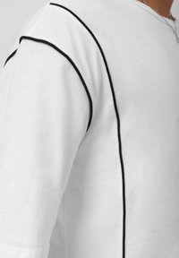 White short-sleeve shirt featuring black piping along the seams, made from a soft cotton material. Close-up shows smooth texture and stitching.
